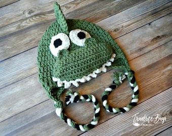 May include: A handmade green crochet hat shaped like a crocodile, featuring white and black eyes, a white-edged mouth, and a fin on top. The hat has braided ties in green, black, and white. The hat is displayed on a wooden surface.