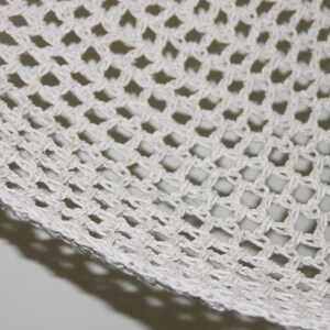 May include: Close-up of a section of white crocheted fabric. The fabric has a grid-like pattern with diamond-shaped openings. The edges of the fabric are visible, showing the woven texture. The background is a light gray.