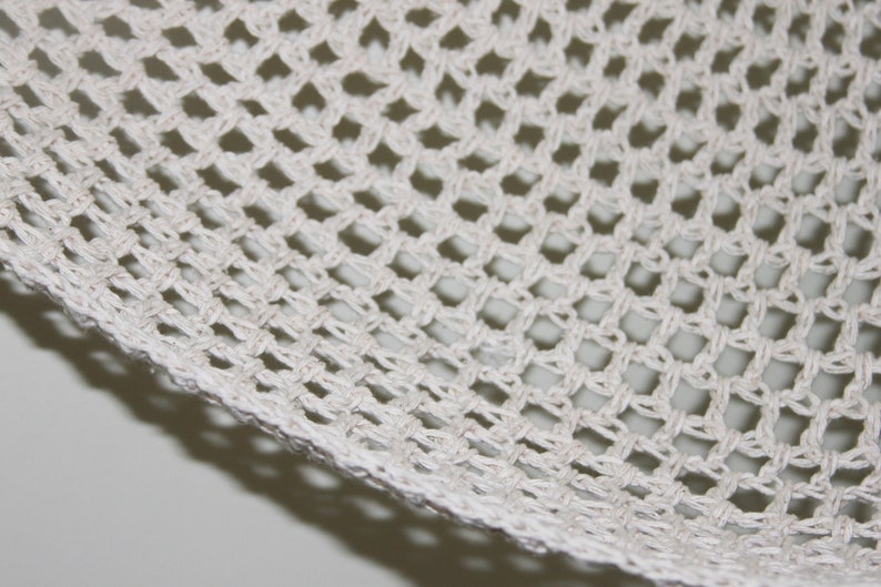 May include: Close-up of a section of white crocheted fabric. The fabric has a grid-like pattern with diamond-shaped openings. The edges of the fabric are visible, showing the woven texture. The background is a light gray.