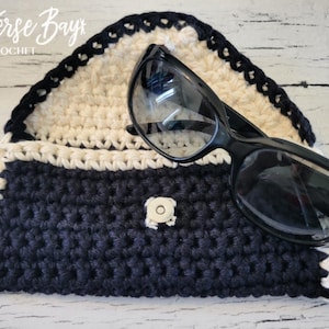 Peut inclure: Un étui à lunettes crocheté bleu marine et crème avec une bordure noire et une fermeture à bouton. Des lunettes noires reposent sur le dessus de l'étui. Le texte "Traverse Bay Crochet" est en haut.