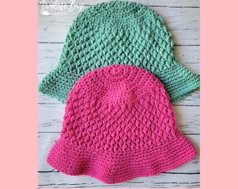 May include: Two handmade crochet bucket hats. One is a vibrant pink, the other is a soft seafoam green. Both hats have a textured stitch pattern and a slightly flared brim. The hats are displayed on a light wood grain surface.