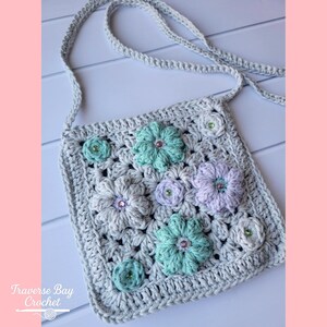 Op de afbeelding: Een gehaakte grijze crossbodytas versierd met kleurrijke bloemenversieringen. De tas heeft een vierkante vorm met een riem en is versierd met gehaakte bloemen in verschillende maten in grijstinten, mintgroen en lavendel. De tekst "Traverse Bay Crochet" is zichtbaar.