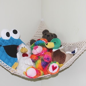May include: A cream-colored, crocheted hammock filled with stuffed animals, including a blue Cookie Monster, a white chicken, and a brown bear. The hammock is suspended in a corner, with a star-shaped decoration above.