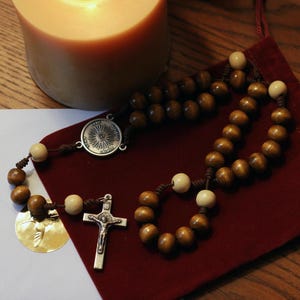 Pu&ograve; includere: Un rosario con grani di legno e un crocifisso d'argento. Il rosario &egrave; appoggiato su un sacchetto di velluto rosso, con una candela accesa sullo sfondo. I grani sono marrone chiaro e il crocifisso raffigura Ges&ugrave;.