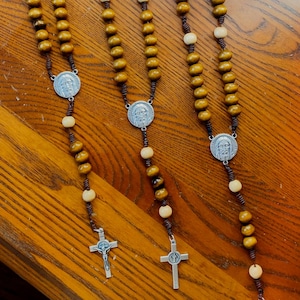Pu&ograve; includere: Diversi rosari con perline di legno nei toni del marrone e del crema sono esposti. Ogni rosario presenta un medaglione d'argento con una figura religiosa e un crocifisso d'argento. I rosari sono disposti su una superficie di legno.