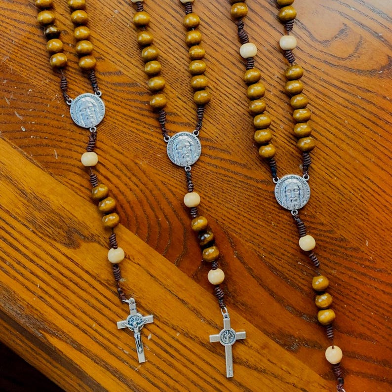 Pu&ograve; includere: Diversi rosari con perline di legno nei toni del marrone e del crema sono esposti. Ogni rosario presenta un medaglione d'argento con una figura religiosa e un crocifisso d'argento. I rosari sono disposti su una superficie di legno.