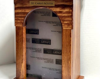 May include: A wooden display case with a dark brown finish. The case features an arched opening and a small plaque that reads "ST. CARLO ACUTIS." The case has a clear, protective front panel.