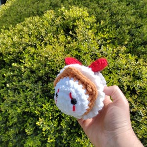 May include: A crocheted plush toy, featuring a character design with white, brown, and red elements. The toy has black eyes and red markings. The plush is held in a hand against a backdrop of green foliage.