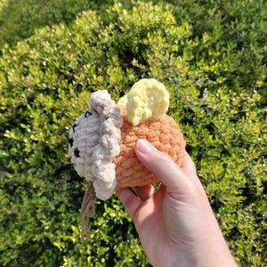 May include: A handmade crochet pumpkin with a tan face, orange body, and yellow stem. The pumpkin has black eyes and a brown string. The pumpkin is held in a hand against a background of green foliage.