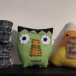 May include: Three Halloween decorations: a green Frankenstein pillow with black eyes and a plaid nose, a gray stone bust of Frankenstein, and a yellow and orange felt candy corn with a brown tag that says "Trick or Treat".