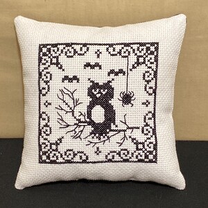 May include: A white pillow with a black embroidered design of a cat sitting on a branch with bats and a spider hanging from a web. The design is framed with a black border.