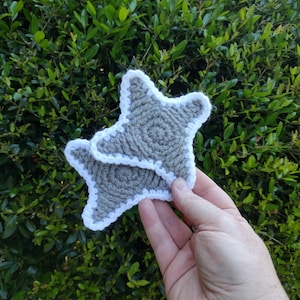 May include: Two gray crocheted star-shaped coasters with white trim are held in a hand against a backdrop of green foliage. The coasters are handmade, featuring a textured design and a contrasting white border, perfect for home decor.