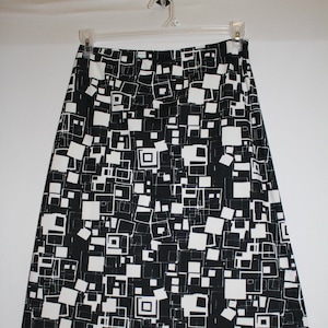 May include: A black and white geometric print skirt hanging on a white hanger. The skirt features a repeating pattern of squares and rectangles in black and white. The skirt is made of a lightweight fabric and appears to be knee-length.