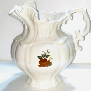 May include: White ceramic pitcher with a ruffled rim and ornate handle. The pitcher features a brown rose floral design on the front. The pitcher is a decorative item.