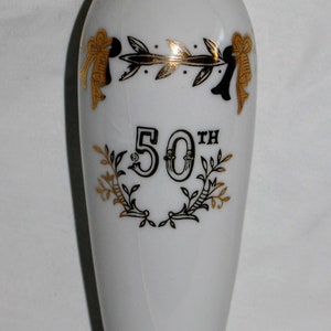 May include: White ceramic vase with gold trim and accents. The vase features a gold and black design with the text "50th" in the center, surrounded by a laurel wreath. Decorative bows are also present.