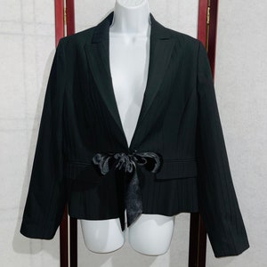 May include: Black blazer with a satin bow closure. The blazer has a notched lapel, two front pockets, and a subtle vertical stripe pattern. The jacket is displayed on a white mannequin.