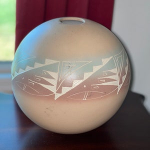 Könnte beinhalten: Eine kugelförmige, beige Keramikvase mit einer kleinen Öffnung oben. Die Vase hat ein geometrisches Design in Weiß und Hellblau in der Mitte. Das Design umfasst Stufenmuster und stilisierte Formen.
