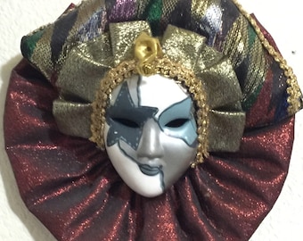 May include: A decorative mask with a harlequin design. The mask features a silver and white face with a star-shaped eye covering and blue accents. It is adorned with a ruffled burgundy collar and a multi-colored, gold-trimmed headpiece.