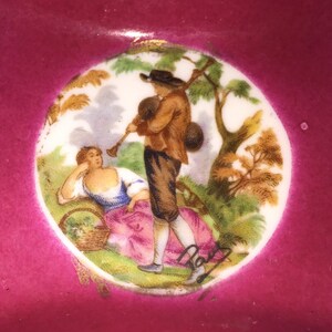 May include: A decorative ceramic plate with a central medallion featuring a pastoral scene. The scene depicts a man playing a wind instrument and a woman reclining, with a basket. The plate has a magenta border and gold accents.