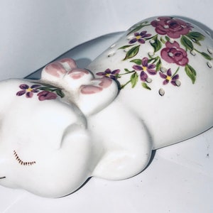 May include: A white ceramic figurine of a sleeping pig, adorned with purple floral accents. The pig has closed eyes and a pink snout and ears. The floral design includes purple flowers and green leaves.