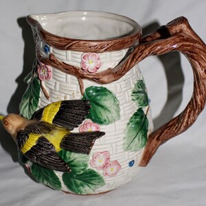 May include: A ceramic pitcher with a woven basket design, adorned with a brown branch handle and a sculpted bird with yellow and black wings. Pink flowers and green leaves decorate the white pitcher.