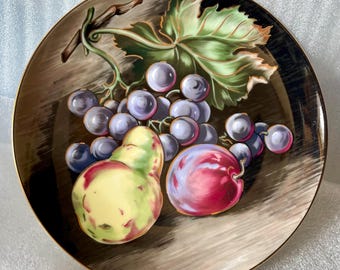May include: A decorative plate featuring a still life of fruit. The plate showcases a cluster of purple grapes, a green and yellow pear, and a red plum. The plate has a gold rim and a brown background.
