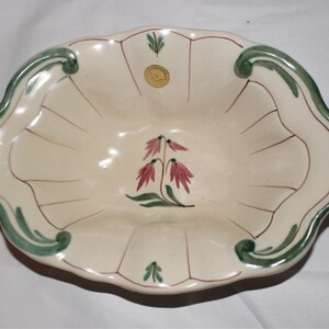 May include: Cream-colored ceramic serving dish with a scalloped edge and a floral design. The dish features a green border, brown accent lines, and a central illustration of pink flowers with green leaves. A small gold sticker is visible.