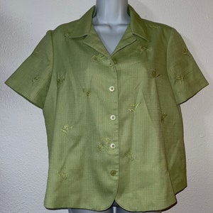 May include: A light green short-sleeved blouse with embroidered floral designs. The blouse has a collared neckline and a button-down front. The fabric has a textured appearance. The floral embroidery is in a matching green color.