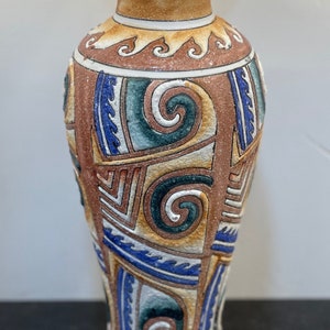 May include: A decorative ceramic vase with a textured surface. The vase features a cream-colored rim and base, with a tan, brown, blue, and green geometric pattern. The design includes swirls and wave-like shapes.