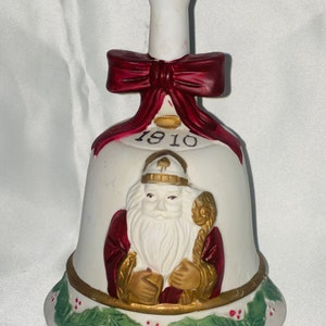 May include: A white ceramic bell featuring a Santa Claus figure with a long white beard, holding a staff. The bell has a red bow and the year "1910" printed on it. The base is decorated with green leaves and red berries.