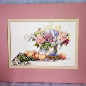 Puede incluir: Pintura a la acuarela de una regadera rebosante de flores coloridas. Melocotones, una peque&ntilde;a cesta de ar&aacute;ndanos y un pa&ntilde;o a cuadros est&aacute;n dispuestos frente a la regadera. La obra est&aacute; enmarcada con un passe-partout rosa.