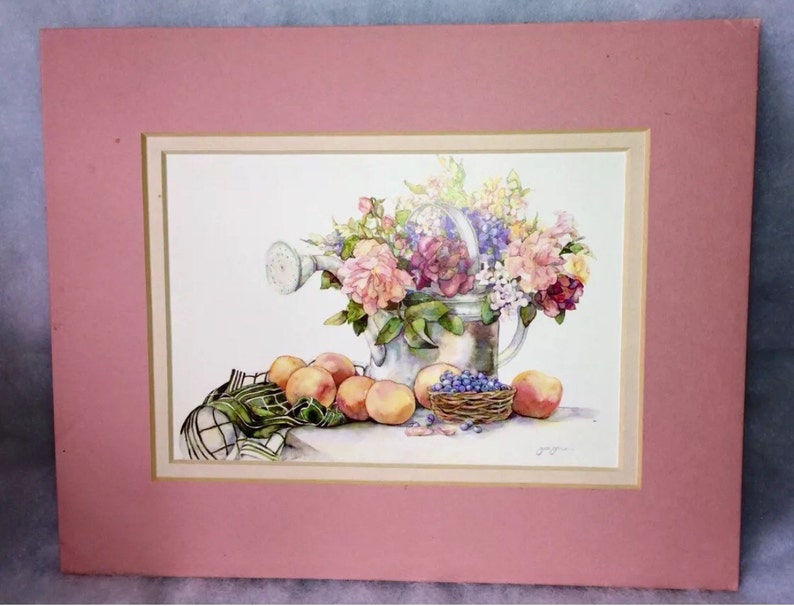 Puede incluir: Pintura a la acuarela de una regadera rebosante de flores coloridas. Melocotones, una peque&ntilde;a cesta de ar&aacute;ndanos y un pa&ntilde;o a cuadros est&aacute;n dispuestos frente a la regadera. La obra est&aacute; enmarcada con un passe-partout rosa.