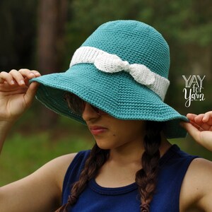 May include: A teal crochet sun hat with a white crocheted ribbon tied in a bow. The hat is being worn by a person with long brown hair. The text "YAY for Yarn" is visible in the background.