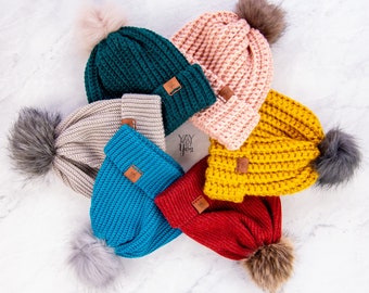 May include: Six crocheted beanies in various colors, including teal, pink, mustard yellow, red, and blue. Each beanie has a fluffy pom-pom on top and a brown leather patch with the text "Yay! Yarn" on it.