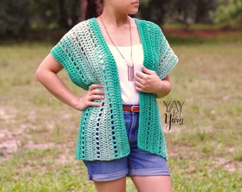May include: A green and white ombre crochet cardigan with short sleeves. The cardigan is open in the front and has a loose fit. The model is wearing the cardigan over a white tank top and denim shorts. The text "YAY for Yarn" is visible in the lower right corner of the image.