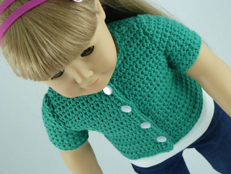 K&ouml;nnte beinhalten: Eine gr&uuml;ne geh&auml;kelte Strickjacke mit wei&szlig;en Kn&ouml;pfen. Die Strickjacke hat kurze &Auml;rmel und einen runden Ausschnitt.