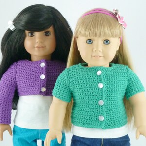 K&ouml;nnte beinhalten: Zwei Puppen tragen geh&auml;kelte Cardigans. Eine Puppe hat schwarze Haare und tr&auml;gt einen lilafarbenen Cardigan. Die andere Puppe hat blonde Haare und tr&auml;gt einen gr&uuml;nen Cardigan.