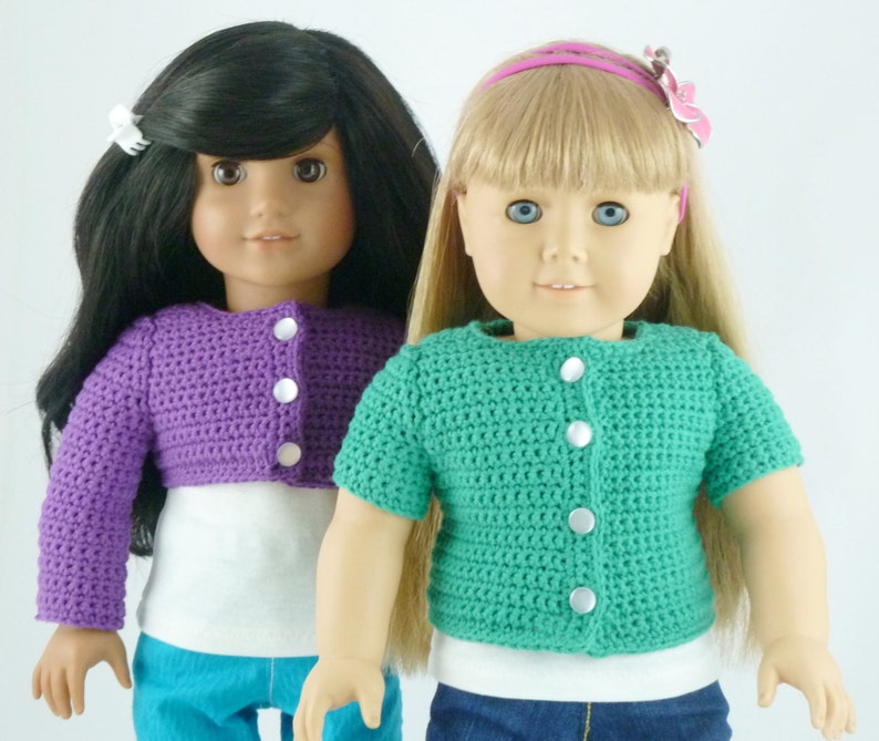 K&ouml;nnte beinhalten: Zwei Puppen tragen geh&auml;kelte Cardigans. Eine Puppe hat schwarze Haare und tr&auml;gt einen lilafarbenen Cardigan. Die andere Puppe hat blonde Haare und tr&auml;gt einen gr&uuml;nen Cardigan.