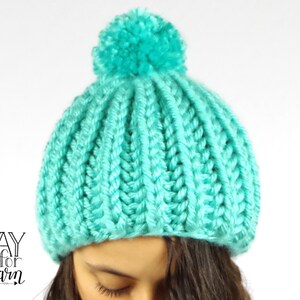 Peut inclure: Un bonnet en tricot turquoise avec un pompon moelleux sur le dessus. Le bonnet est fait avec un fil épais et a une texture côtelée.
