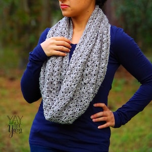 May include: A gray crocheted infinity scarf with a delicate openwork pattern. The scarf is worn by a person with a blue long-sleeved shirt.