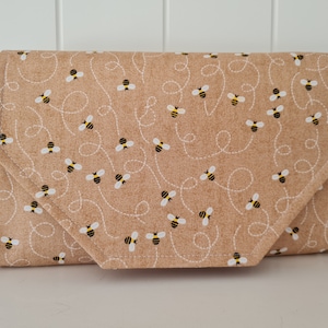 May include: A beige fabric clutch with a pattern of small yellow bees flying in a line. The clutch has a flap closure and a zippered pocket on the side.