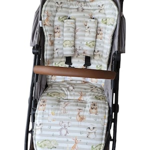 Könnte beinhalten: Ein Kinderwagen-Sitzbezug mit einem grau-weiß gestreiften Hintergrund, auf dem verschiedene australische Tiere abgebildet sind, darunter Koalas, Kängurus und Krokodile. Der Bezug hat zwei gepolsterte Gurte.