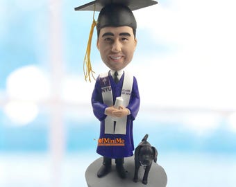 Könnte beinhalten: Eine personalisierte Wackelkopf-Figur eines Absolventen in einem violetten akademischen Talar, einer weißen Stola mit der Aufschrift NYU und Class of 2018 sowie einem schwarzen Doktorhut mit gelber Quaste. Die Figur hält ein weißes Diplom und einen schwarzen Hund.