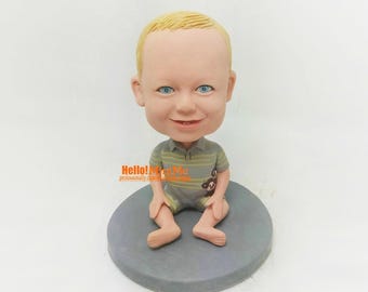 May include: A custom-made figurine of a young child with blonde hair, wearing a gray and yellow striped shirt and gray shorts. The figurine is sitting on a gray base.