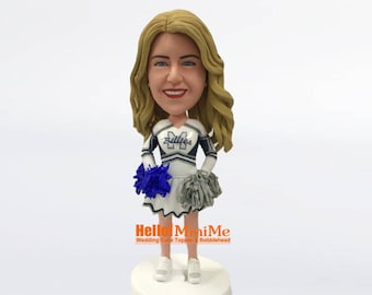 May include: A custom bobblehead figurine of a woman wearing a white and blue cheerleading uniform with a blue and white pom-pom. The uniform has the word "Bullies" on the front. The bobblehead is on a white base.