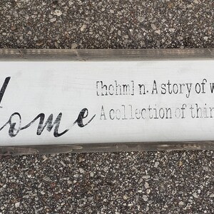 May include: A white wooden sign with a black painted inscription that reads "Home [hohm] n. A story of who we are. A collection of things we love."