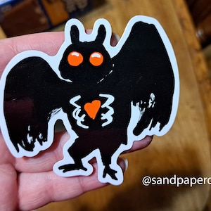 May include: Black and white sticker of a cartoon moth with orange eyes and a red heart on its chest. The sticker is on a white background and has the text "@sandpaperdaisy" at the bottom.