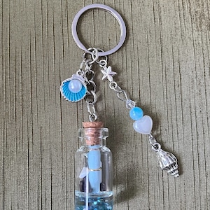 May include: A silver keychain with a small glass bottle filled with blue glitter, a rolled up piece of paper, and a small seashell. The keychain also has a silver star charm, a blue bead, a white heart charm, and a silver seashell charm.