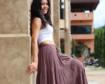 May include: A woman wearing a white tank top and a long, tiered, mauve skirt. She is standing next to a textured wall and smiling.