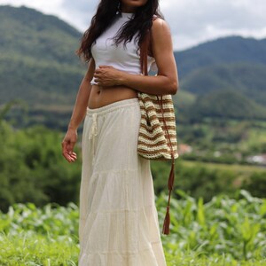K&ouml;nnte beinhalten: Eine Frau tr&auml;gt ein wei&szlig;es Crop-Top und einen wei&szlig;en, mehrlagigen Maxirock mit einer braunen und wei&szlig;en geh&auml;kelten Tasche. Sie steht auf einem Feld mit gr&uuml;nem Gras und Bergen im Hintergrund.